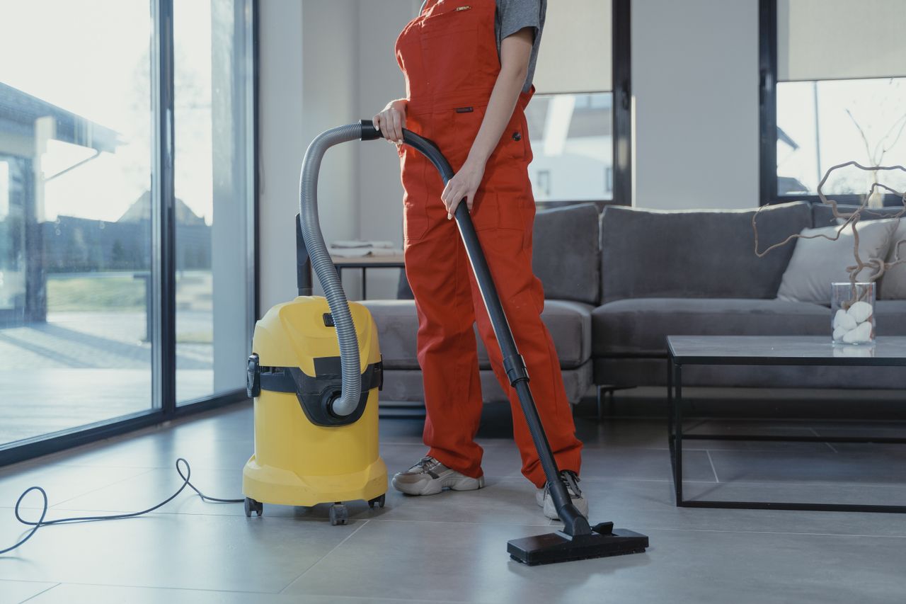 Un professionnel du nettoyage en combinaison rouge aspire un salon moderne avec un aspirateur jaune.